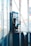 A vintage-style telephone booth housing a blue and silver payphone is situated in a sunlit, slightly weathered urban setting with clear glass walls.