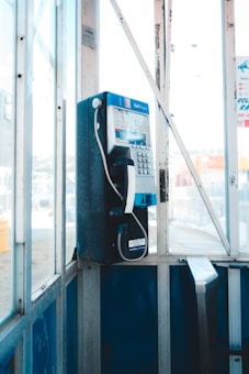 A vintage-style telephone booth housing a blue and silver payphone is situated in a sunlit, slightly weathered urban setting with clear glass walls.