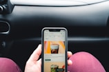 A person is holding a smartphone displaying a coffee shop app, showing an advertisement for cold brew coffee. The phone is held in front of a car dashboard. The background includes part of a steering wheel and the car's interior.