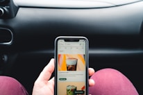 A person is holding a smartphone displaying a coffee shop app, showing an advertisement for cold brew coffee. The phone is held in front of a car dashboard. The background includes part of a steering wheel and the car's interior.