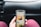 A person is holding a smartphone displaying a coffee shop app, showing an advertisement for cold brew coffee. The phone is held in front of a car dashboard. The background includes part of a steering wheel and the car's interior.
