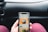 A person is holding a smartphone displaying a coffee shop app, showing an advertisement for cold brew coffee. The phone is held in front of a car dashboard. The background includes part of a steering wheel and the car's interior.