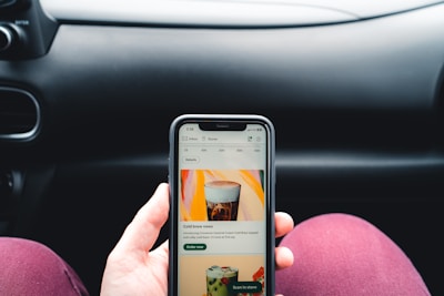 A person is holding a smartphone displaying a coffee shop app, showing an advertisement for cold brew coffee. The phone is held in front of a car dashboard. The background includes part of a steering wheel and the car's interior.