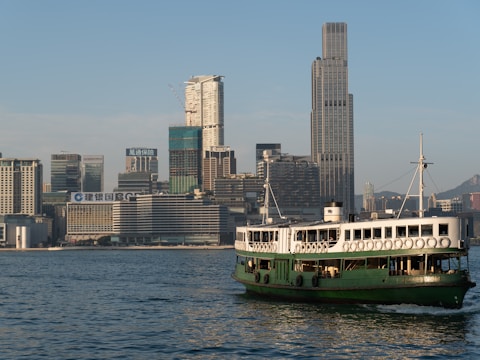 A green ferry sails across a body of water with a city skyline in the background, featuring modern skyscrapers and high-rise buildings under a clear blue sky.