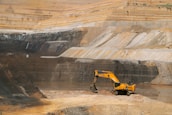 A large open-pit mine with a yellow excavator positioned at the center, surrounded by layered rock formations exhibiting varied shades of brown and gray. The broad expanse of the mining area showcases distinct strata and erosion patterns carved into the earth.