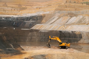 A large open-pit mine with a yellow excavator positioned at the center, surrounded by layered rock formations exhibiting varied shades of brown and gray. The broad expanse of the mining area showcases distinct strata and erosion patterns carved into the earth.