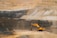 A large open-pit mine with a yellow excavator positioned at the center, surrounded by layered rock formations exhibiting varied shades of brown and gray. The broad expanse of the mining area showcases distinct strata and erosion patterns carved into the earth.