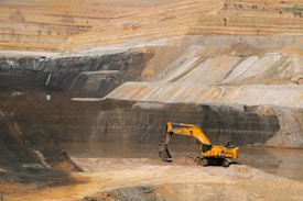 A large open-pit mine with a yellow excavator positioned at the center, surrounded by layered rock formations exhibiting varied shades of brown and gray. The broad expanse of the mining area showcases distinct strata and erosion patterns carved into the earth.