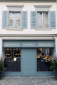 A storefront with a teal-blue facade featuring the name 'Beatrice Rossi Schmuck' written in gold letters. The building has two large windows on either side of a closed, teal-blue roller shutter, and the windows display various merchandise inside. Above the storefront are two pairs of grey shuttered windows on the upper level. There are small trees placed on both sides of the entrance.