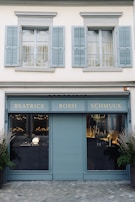 A storefront with a teal-blue facade featuring the name 'Beatrice Rossi Schmuck' written in gold letters. The building has two large windows on either side of a closed, teal-blue roller shutter, and the windows display various merchandise inside. Above the storefront are two pairs of grey shuttered windows on the upper level. There are small trees placed on both sides of the entrance.