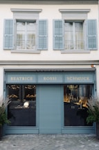 A storefront with a teal-blue facade featuring the name 'Beatrice Rossi Schmuck' written in gold letters. The building has two large windows on either side of a closed, teal-blue roller shutter, and the windows display various merchandise inside. Above the storefront are two pairs of grey shuttered windows on the upper level. There are small trees placed on both sides of the entrance.