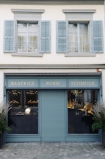 A storefront with a teal-blue facade featuring the name 'Beatrice Rossi Schmuck' written in gold letters. The building has two large windows on either side of a closed, teal-blue roller shutter, and the windows display various merchandise inside. Above the storefront are two pairs of grey shuttered windows on the upper level. There are small trees placed on both sides of the entrance.