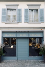 A storefront with a teal-blue facade featuring the name 'Beatrice Rossi Schmuck' written in gold letters. The building has two large windows on either side of a closed, teal-blue roller shutter, and the windows display various merchandise inside. Above the storefront are two pairs of grey shuttered windows on the upper level. There are small trees placed on both sides of the entrance.