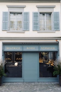 A storefront with a teal-blue facade featuring the name 'Beatrice Rossi Schmuck' written in gold letters. The building has two large windows on either side of a closed, teal-blue roller shutter, and the windows display various merchandise inside. Above the storefront are two pairs of grey shuttered windows on the upper level. There are small trees placed on both sides of the entrance.
