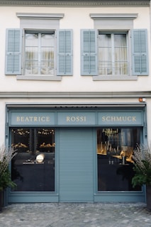 A storefront with a teal-blue facade featuring the name 'Beatrice Rossi Schmuck' written in gold letters. The building has two large windows on either side of a closed, teal-blue roller shutter, and the windows display various merchandise inside. Above the storefront are two pairs of grey shuttered windows on the upper level. There are small trees placed on both sides of the entrance.