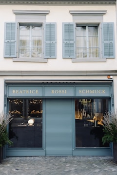 A storefront with a teal-blue facade featuring the name 'Beatrice Rossi Schmuck' written in gold letters. The building has two large windows on either side of a closed, teal-blue roller shutter, and the windows display various merchandise inside. Above the storefront are two pairs of grey shuttered windows on the upper level. There are small trees placed on both sides of the entrance.