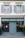 A storefront with a teal-blue facade featuring the name 'Beatrice Rossi Schmuck' written in gold letters. The building has two large windows on either side of a closed, teal-blue roller shutter, and the windows display various merchandise inside. Above the storefront are two pairs of grey shuttered windows on the upper level. There are small trees placed on both sides of the entrance.