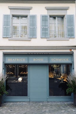 A storefront with a teal-blue facade featuring the name 'Beatrice Rossi Schmuck' written in gold letters. The building has two large windows on either side of a closed, teal-blue roller shutter, and the windows display various merchandise inside. Above the storefront are two pairs of grey shuttered windows on the upper level. There are small trees placed on both sides of the entrance.