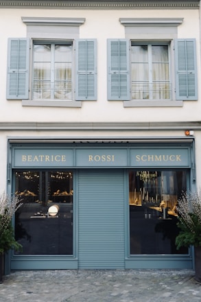 A storefront with a teal-blue facade featuring the name 'Beatrice Rossi Schmuck' written in gold letters. The building has two large windows on either side of a closed, teal-blue roller shutter, and the windows display various merchandise inside. Above the storefront are two pairs of grey shuttered windows on the upper level. There are small trees placed on both sides of the entrance.