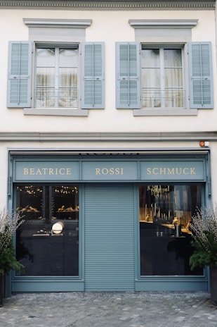 A storefront with a teal-blue facade featuring the name 'Beatrice Rossi Schmuck' written in gold letters. The building has two large windows on either side of a closed, teal-blue roller shutter, and the windows display various merchandise inside. Above the storefront are two pairs of grey shuttered windows on the upper level. There are small trees placed on both sides of the entrance.