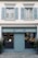 A storefront with a teal-blue facade featuring the name 'Beatrice Rossi Schmuck' written in gold letters. The building has two large windows on either side of a closed, teal-blue roller shutter, and the windows display various merchandise inside. Above the storefront are two pairs of grey shuttered windows on the upper level. There are small trees placed on both sides of the entrance.