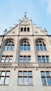 A detailed architectural image of a multi-story building facade featuring large arched windows with wooden frames. The structure includes ornate sculptural elements and decorative stonework. The top section of the building showcases smaller arched openings and a pinnacle with a small spire.