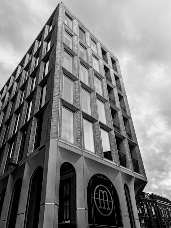 A tall, multi-story building with a modern architectural design featuring large rectangular windows and brickwork. The building has an angular appearance, rising into a cloudy sky. The lower part of the structure includes large arched entrances, and a circular logo with the letter 'm' is visible on one of the arches. The surrounding area shows other buildings partially in view.