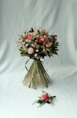 A beautifully arranged bouquet consisting of various flowers and greenery. The bouquet features pink roses, white daisies, and other floral elements, bound together neatly with long green leaves. It is accompanied by a smaller floral arrangement placed on a light grey background.