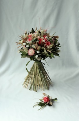 A beautifully arranged bouquet consisting of various flowers and greenery. The bouquet features pink roses, white daisies, and other floral elements, bound together neatly with long green leaves. It is accompanied by a smaller floral arrangement placed on a light grey background.