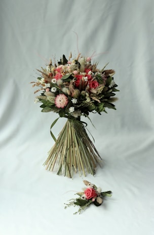 A beautifully arranged bouquet consisting of various flowers and greenery. The bouquet features pink roses, white daisies, and other floral elements, bound together neatly with long green leaves. It is accompanied by a smaller floral arrangement placed on a light grey background.