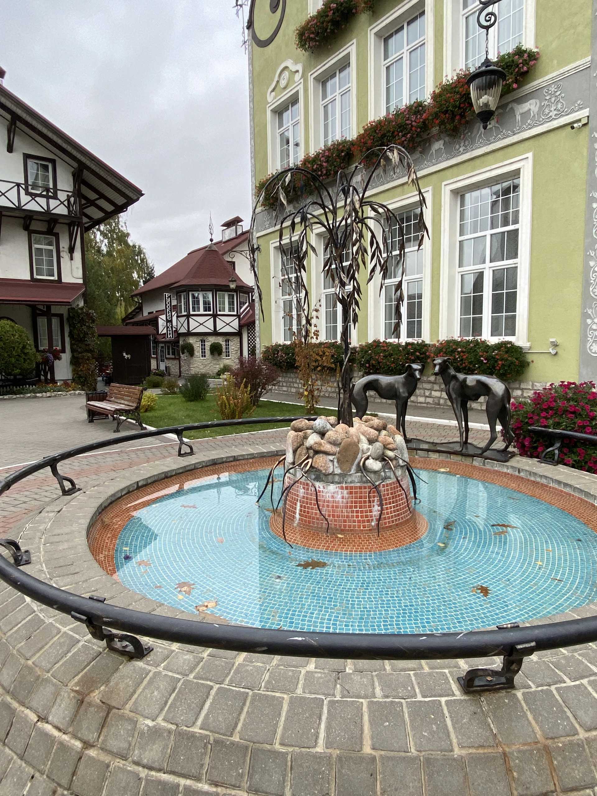 A charming courtyard features a decorative fountain with a tiled basin and stone centerpieces, surrounded by landscaped gardens. The backdrop includes quaint buildings with large windows, ornate designs, and flower boxes under overcast skies.