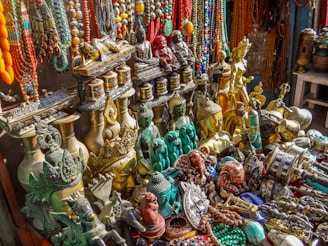 An assortment of colorful and ornate statues, beads, and trinkets, including various Buddha and deity figures, intricately designed vases, and prayer wheels. The items are made of materials like brass and are richly detailed, showing craftsmanship. Strings of vibrant beads hang in the background, adding to the array of colors and textures.