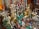An assortment of colorful and ornate statues, beads, and trinkets, including various Buddha and deity figures, intricately designed vases, and prayer wheels. The items are made of materials like brass and are richly detailed, showing craftsmanship. Strings of vibrant beads hang in the background, adding to the array of colors and textures.