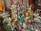 An assortment of colorful and ornate statues, beads, and trinkets, including various Buddha and deity figures, intricately designed vases, and prayer wheels. The items are made of materials like brass and are richly detailed, showing craftsmanship. Strings of vibrant beads hang in the background, adding to the array of colors and textures.