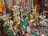 An assortment of colorful and ornate statues, beads, and trinkets, including various Buddha and deity figures, intricately designed vases, and prayer wheels. The items are made of materials like brass and are richly detailed, showing craftsmanship. Strings of vibrant beads hang in the background, adding to the array of colors and textures.