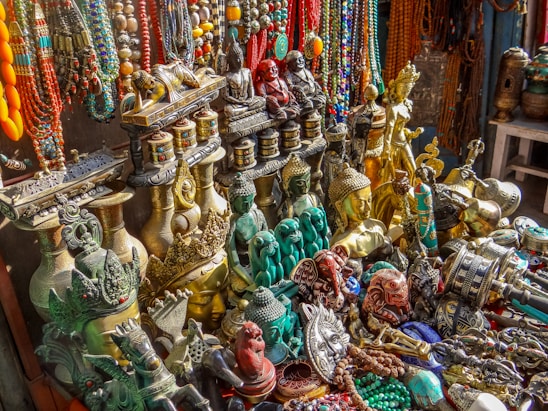 An assortment of colorful and ornate statues, beads, and trinkets, including various Buddha and deity figures, intricately designed vases, and prayer wheels. The items are made of materials like brass and are richly detailed, showing craftsmanship. Strings of vibrant beads hang in the background, adding to the array of colors and textures.