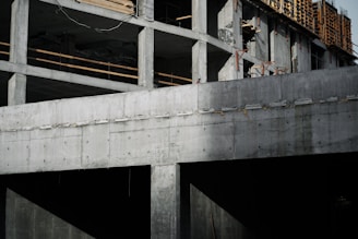 A partially constructed concrete building with visible structural columns and beams. The building features exposed rebar and unfinished surfaces, suggesting an early stage of construction. Sunlight casts shadows across the surfaces, enhancing the textures of the concrete.
