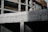 A partially constructed concrete building with visible structural columns and beams. The building features exposed rebar and unfinished surfaces, suggesting an early stage of construction. Sunlight casts shadows across the surfaces, enhancing the textures of the concrete.