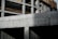 A partially constructed concrete building with visible structural columns and beams. The building features exposed rebar and unfinished surfaces, suggesting an early stage of construction. Sunlight casts shadows across the surfaces, enhancing the textures of the concrete.