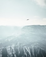 A solitary drone flies high above snow-covered mountains under a vast, pale sky. The landscape is expansive, with rugged peaks and ridges rising from the earth, covered in patches of snow and frost. The atmosphere is calm and serene, emphasizing the contrast between the small, dark drone and the immense natural backdrop.