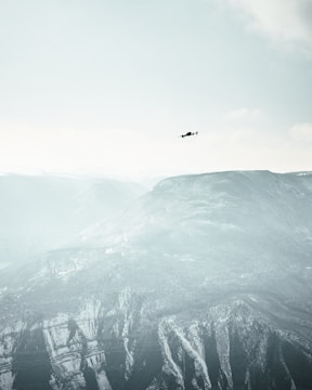 A solitary drone flies high above snow-covered mountains under a vast, pale sky. The landscape is expansive, with rugged peaks and ridges rising from the earth, covered in patches of snow and frost. The atmosphere is calm and serene, emphasizing the contrast between the small, dark drone and the immense natural backdrop.