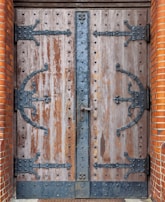 A large, double wooden door with heavy black wrought iron hinges and fittings. The wood appears aged and weathered with visible grain and imperfections. The ironwork includes intricate designs and rivets, with a vertical iron strip running down the center where the doors meet, featuring a central lock. The edges of the doorway are framed with red-orange brickwork.