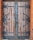 A large, double wooden door with heavy black wrought iron hinges and fittings. The wood appears aged and weathered with visible grain and imperfections. The ironwork includes intricate designs and rivets, with a vertical iron strip running down the center where the doors meet, featuring a central lock. The edges of the doorway are framed with red-orange brickwork.