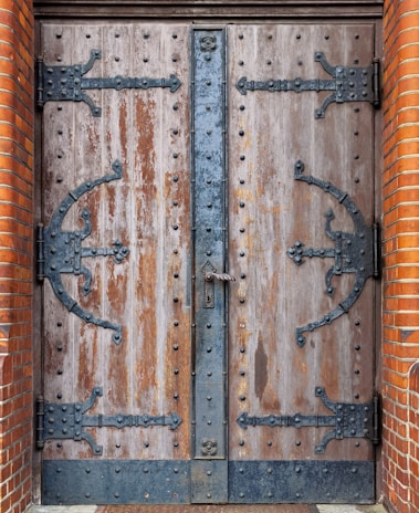 A large, double wooden door with heavy black wrought iron hinges and fittings. The wood appears aged and weathered with visible grain and imperfections. The ironwork includes intricate designs and rivets, with a vertical iron strip running down the center where the doors meet, featuring a central lock. The edges of the doorway are framed with red-orange brickwork.