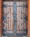 A large, double wooden door with heavy black wrought iron hinges and fittings. The wood appears aged and weathered with visible grain and imperfections. The ironwork includes intricate designs and rivets, with a vertical iron strip running down the center where the doors meet, featuring a central lock. The edges of the doorway are framed with red-orange brickwork.