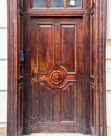 A richly carved wooden door with intricate detailing and a prominent central circular pattern. The wood displays natural grain patterns and a reddish-brown finish. The door is surrounded by a stone frame, and there is a small black mailbox or intercom on the left side.