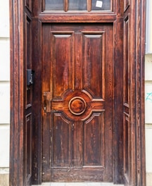 A richly carved wooden door with intricate detailing and a prominent central circular pattern. The wood displays natural grain patterns and a reddish-brown finish. The door is surrounded by a stone frame, and there is a small black mailbox or intercom on the left side.