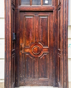 A richly carved wooden door with intricate detailing and a prominent central circular pattern. The wood displays natural grain patterns and a reddish-brown finish. The door is surrounded by a stone frame, and there is a small black mailbox or intercom on the left side.