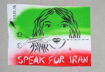 A stenciled image featuring the face of a woman with scissors cutting through a line across her face. The background is painted in green, white, and red colors. The text 'SPEAK FOR IRAN' is prominently displayed in white across the red section.