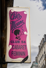 A vertical sign displays the name 'Chez Moune' with an elegant script font against a pink background. Below, the text 'Club 54 Cabaret Feminin' is written in bold, black letters. An illustration of a woman's profile, styled in a vintage silhouette, is depicted as well. The sign is mounted on the exterior of a building against a backdrop of clouds and neighboring structures.